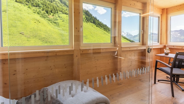 Une pièce de cabane en bois dotée de grandes fenêtres offrant une vue sur une colline verdoyante et des montagnes.
