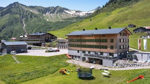 Un bâtiment moderne doté d’un toit plat et de nombreuses fenêtres, entouré d’un paysage verdoyant et avec des montagnes en arrière-plan.