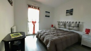 A bedroom with a bed, a nightstand, a table, and a window with curtains. - Flat for 4 , 150m from the beach in Sion (Saint-Hilaire-de-Riez)