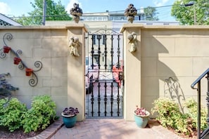 Stately entrance with ornate metal gate and lion statues, surrounded by potted plants and a brick pathway.