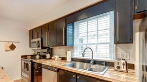 A modern kitchen with dark cabinets, stainless steel appliances, a double sink, and a window with blinds. - Spacious Garden Unit on Historic Jones Street! (Savannah)