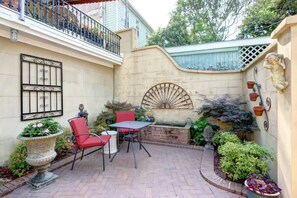 patio area with a brick floor, small table, and two chairs, surrounded by lush greenery and a decorative wall with a sunburst pattern.