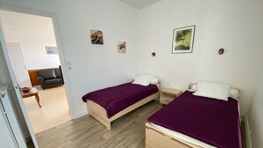 A bedroom with two beds, wooden flooring, and a door leading to a living area with a sofa and a table. - MOUETTES 3 - Apartment with sea view between Carnac and Trinité-sur-Mer - T356 (La Trinité-sur-Mer)