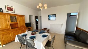 A dining area with a table set for four, a wooden cabinet with glass doors, a black leather sofa, and a ceiling light fixture. - MOUETTES 3 - Apartment with sea view between Carnac and Trinité-sur-Mer - T356 (La Trinité-sur-Mer)
