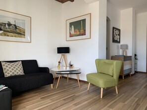 A modern living room with a black sofa, a green armchair, a round table with a lamp, and framed artwork on the walls.