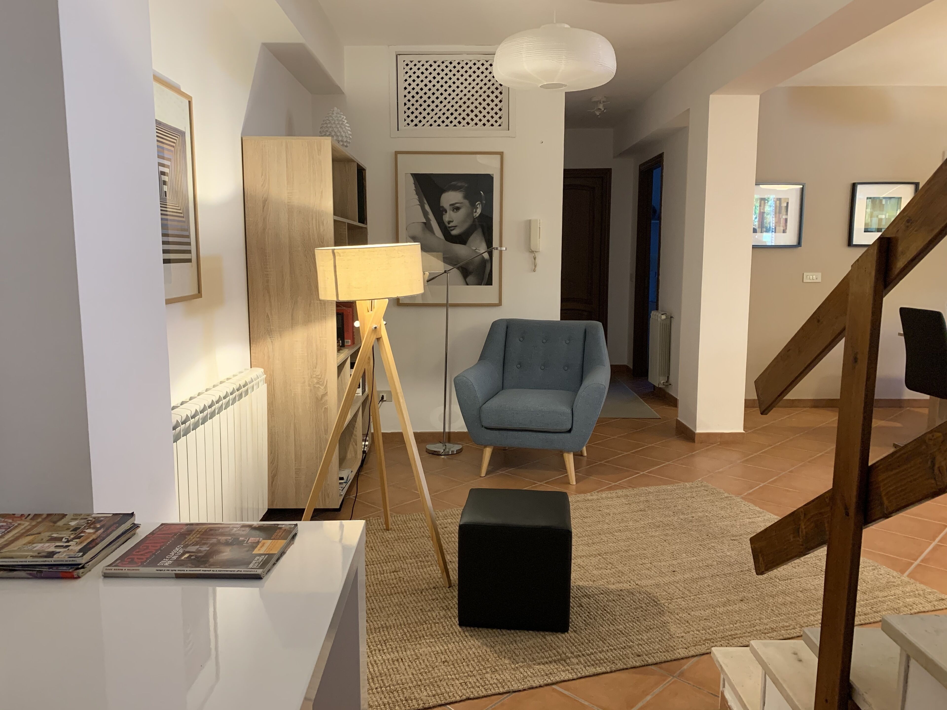 A modern living room with a wooden floor, a blue armchair, a standing lamp, a black ottoman, and a white coffee table.