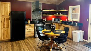 Een kleine keuken met houten kastjes, een zwarte koelkast, een fornuis en een magnetron. Er staat een eettafel met stoelen en een kleine afvalbak.