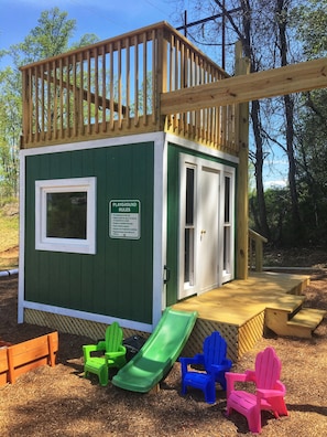 Une petite maison de jeu en bois, dotée d’un toboggan, d’une porte et d’un panneau indiquant les règles à respecter sur l’aire de jeux.