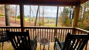 Een houten terras met twee stoelen en een kleine tafel, met uitzicht op een fraai landschap met bomen en een huis in de verte.