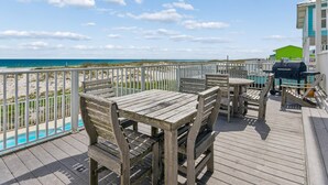 Eine Holzterrasse mit Blick auf Strand und Meer, ein Grill und ein grünes Gebäude im Hintergrund.
