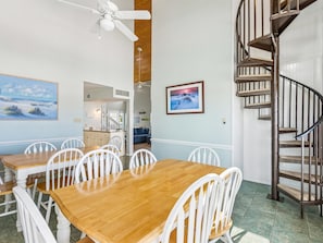 Un coin repas avec une table en bois et des chaises blanches, un ventilateur de plafond, un escalier en colimaçon et un tableau accroché au mur.