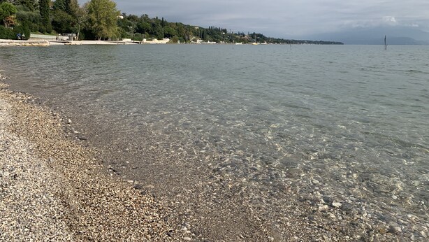 A pebbled beach with clear water, trees, and buildings in the background. - Hibiscus apartment | Garda Lake & Golf (Padenghe Sul Garda)