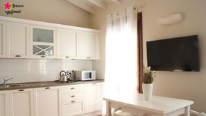 A modern kitchen with white cabinets, a microwave, and a kettle on the counter. There is a dining table with chairs and a flat-screen TV mounted on the wall. - Hibiscus apartment | Garda Lake & Golf (Padenghe Sul Garda)