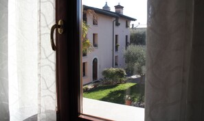 Blick durch ein Fenster mit Vorhang, der ein Gebäude mit zahlreichen Fenstern und einem Garten zeigt.