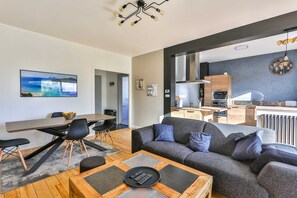 Uma sala de estar moderna com um sofá cinza, uma mesa de centro de madeira e um espaço de jantar com mesa e cadeiras de madeira. Uma cozinha com painel atrás do fogão cinza e armários de madeira é visível ao fundo.