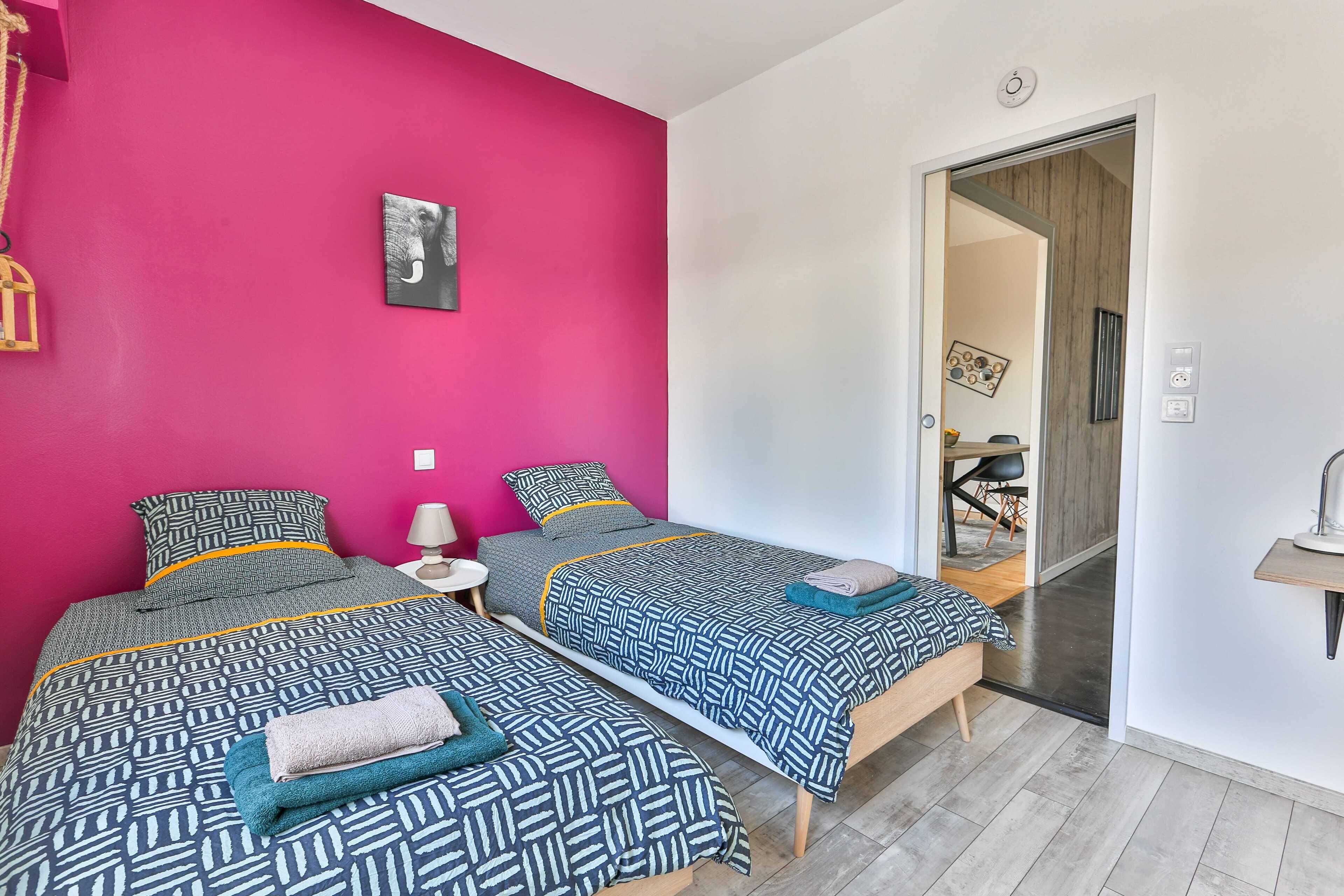 A room with two beds, a pink wall, a door leading to another room, and a small bedside table with a lamp.