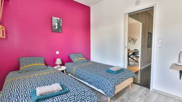 A room with two beds, a pink wall, a door leading to another room, and a small bedside table with a lamp.
