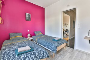 A room with two beds, a pink wall, a door leading to another room, and a small bedside table with a lamp.