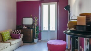 Een woonkamer met een bank, een dressoir, een potplant, een roze pouf en een tafel met kaarsen en een lamp.