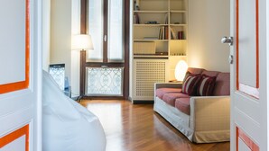 Un pasillo con suelo de madera, un sillón inflable blanco, un sofá con cojín a rayas y una estantería con libros y una lámpara.