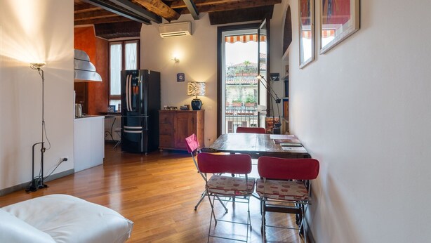 Una habitación amplia con suelo de madera, una mesa de comedor con sillas, una zona de cocina con refrigerador y un ventanal que ofrece vistas al exterior.