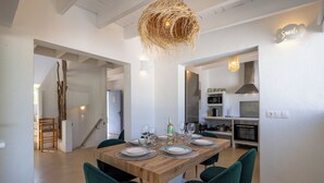 A dining area with a wooden table set for four, surrounded by green chairs, a hanging light fixture, and a kitchen in the background. - Villa Clara Ibiza with pool (Santa Eulària des Riu)