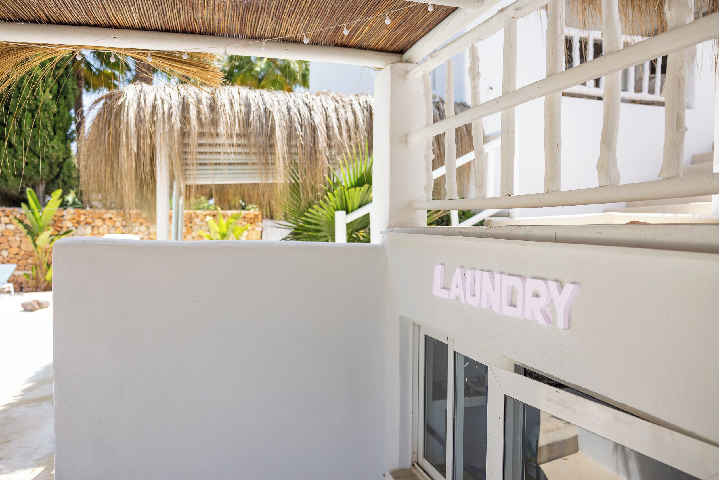 Un edificio blanco con un letrero que dice 'Laundry', techo de paja y un muro de piedra al fondo.