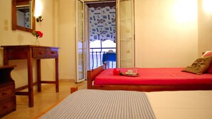 Une chambre avec un lit rouge, une commode en bois, un miroir et une vue sur l’extérieur par une porte ouverte.