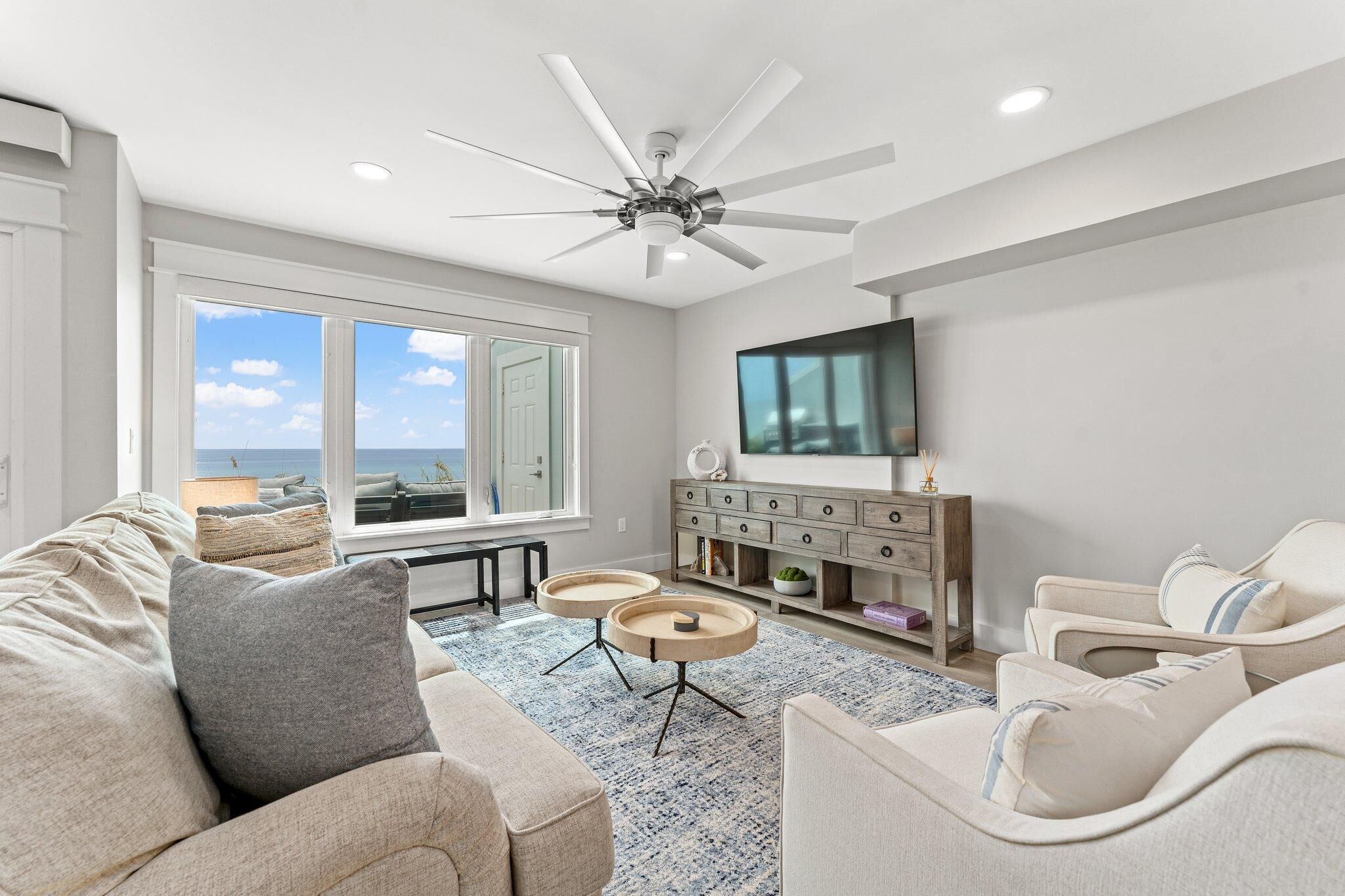 A modern living room with a ceiling fan, large windows, a flat-screen TV, and a cozy seating area.