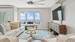 A modern living room with a ceiling fan, large windows with a view of the ocean, a TV mounted on the wall, and a wooden cabinet. - Stunning Gulf Front Townhouse! Private beach, beach chairs, wagon, and cooler! (Santa Rosa Beach)