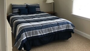 Un dormitorio con cama, ventilador de techo, ventana con persianas y mesita de noche con lámpara.