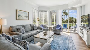 A spacious living room with a grey sofa, a coffee table, and a TV on a wooden cabinet. There is a balcony with a view of palm trees and a pool. - Enjoy Comfort & Peace w/ Balcony and Amenities (Hilton Head Island)