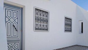 A white building with a patterned door, two windows with decorative iron grilles, and a tiled entrance area.