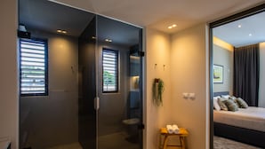 A modern hotel room with a glass partition, a small wooden bench with towels, a bed with pillows, and a plant.