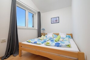 Ein Einzelbett mit Holzrahmen und einer weißen Matratze, ein Nachttisch mit Lampe, ein Fenster mit Vorhängen und ein gerahmtes Bild an der Wand.