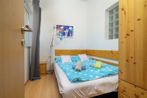 Quarto com cabeceira de madeira, cama com roupa de cama azul com bolinhas, abajur e janela com persianas.