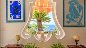 A room with a large window, framed artwork, and two side tables with lamps. - Villa Faraglioni Capri (Capri)