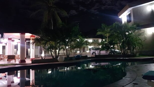 A nighttime pool area with palm trees, a covered seating area, and a building in the background. - Naf Apartments - Mtwara (Mtwara)