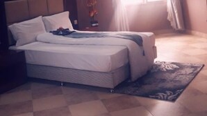 A bedroom with a large bed, a nightstand, a decorative vase, and a window with sheer curtains. - Naf Apartments - Mtwara (Mtwara)