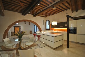 A modern kitchen with a central island, wooden beams on the ceiling, and a large arched doorway. - Il Frantoio - Charming Loft in the old town (Radicondoli)