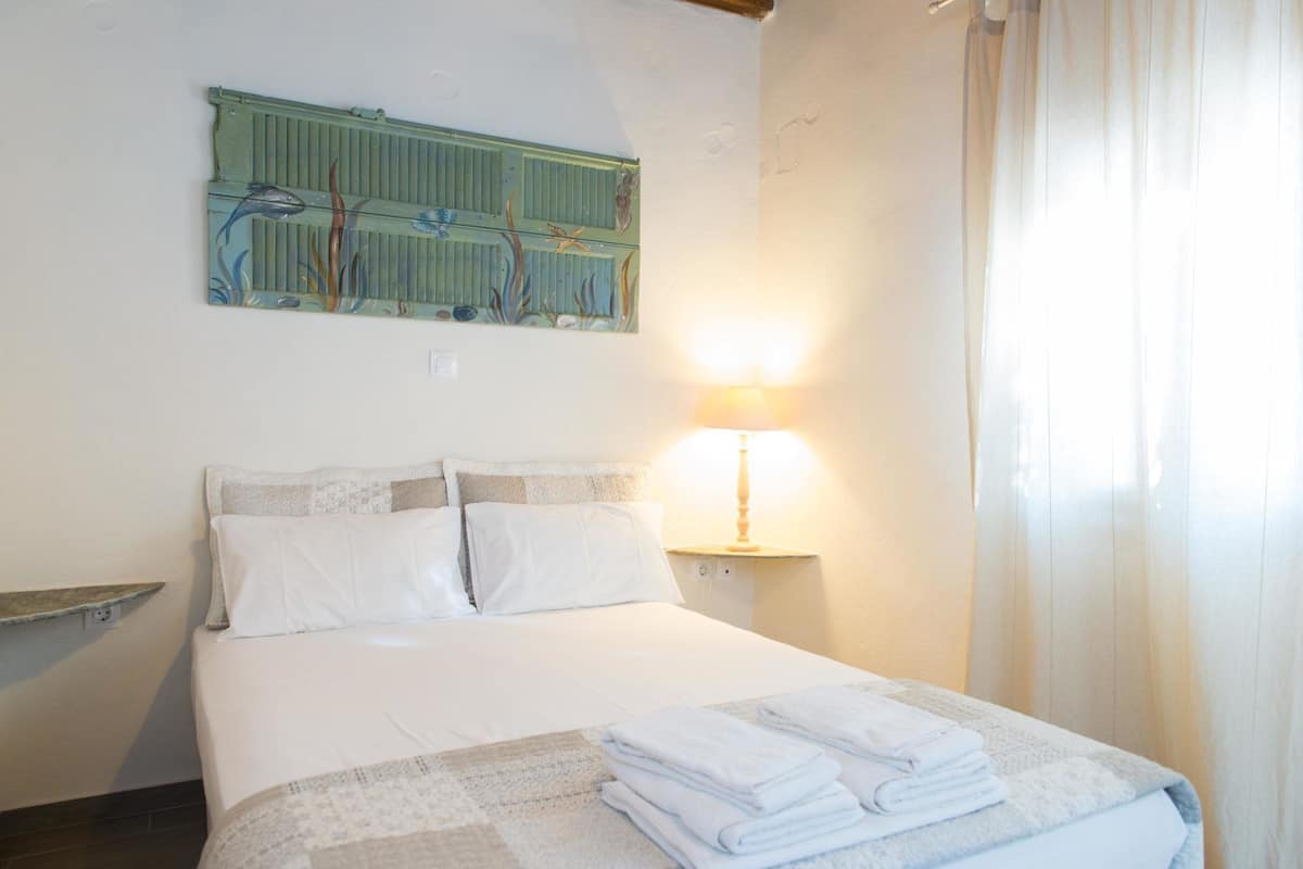 A neatly made bed with white linens and a folded towel on the bed. There is a wall-mounted artwork depicting marine life above the bed. A bedside lamp is on to the right of the bed.