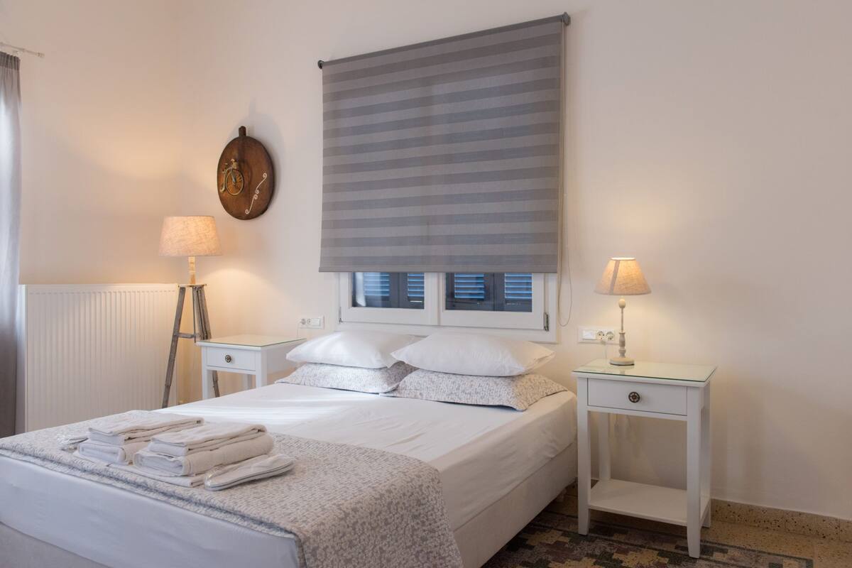 A hotel room with a bed, two bedside tables, a lamp, a window with blinds, and a wall clock.