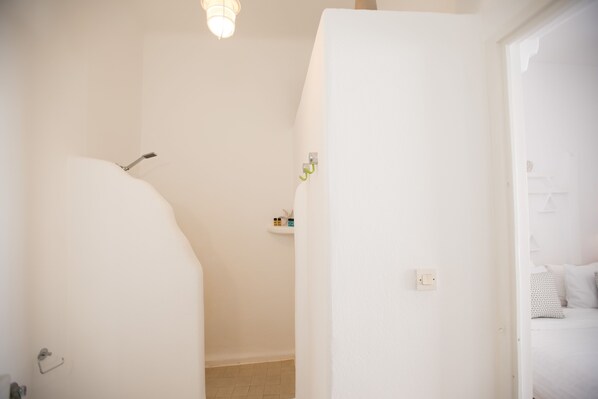 A white bathroom with a shower, a small shelf with toiletries, and a bed with white bedding in the adjacent room. - Two bedroom complex with veranda (Mikonos)