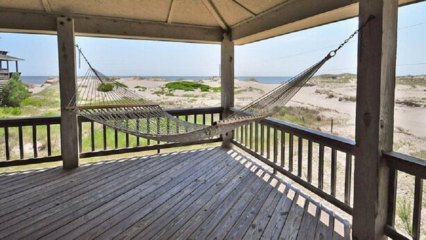 Une terrasse en bois avec un hamac, donnant sur une plage bordée de dunes et sous un ciel dégagé.