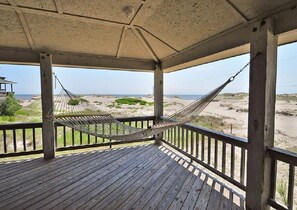 A wooden deck with a hammock, overlooking a beach with dunes and a clear sky.