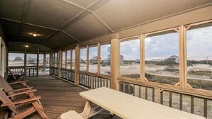 Une terrasse extérieure couverte, dotée de bancs en bois et d’une table de pique-nique, qui offre une vue sur une plage bordée de dunes et sur des bâtiments situés au loin.
