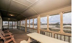 Une terrasse extérieure couverte, dotée de bancs en bois et d’une table de pique-nique, qui offre une vue sur une plage bordée de dunes et sur des bâtiments situés au loin.