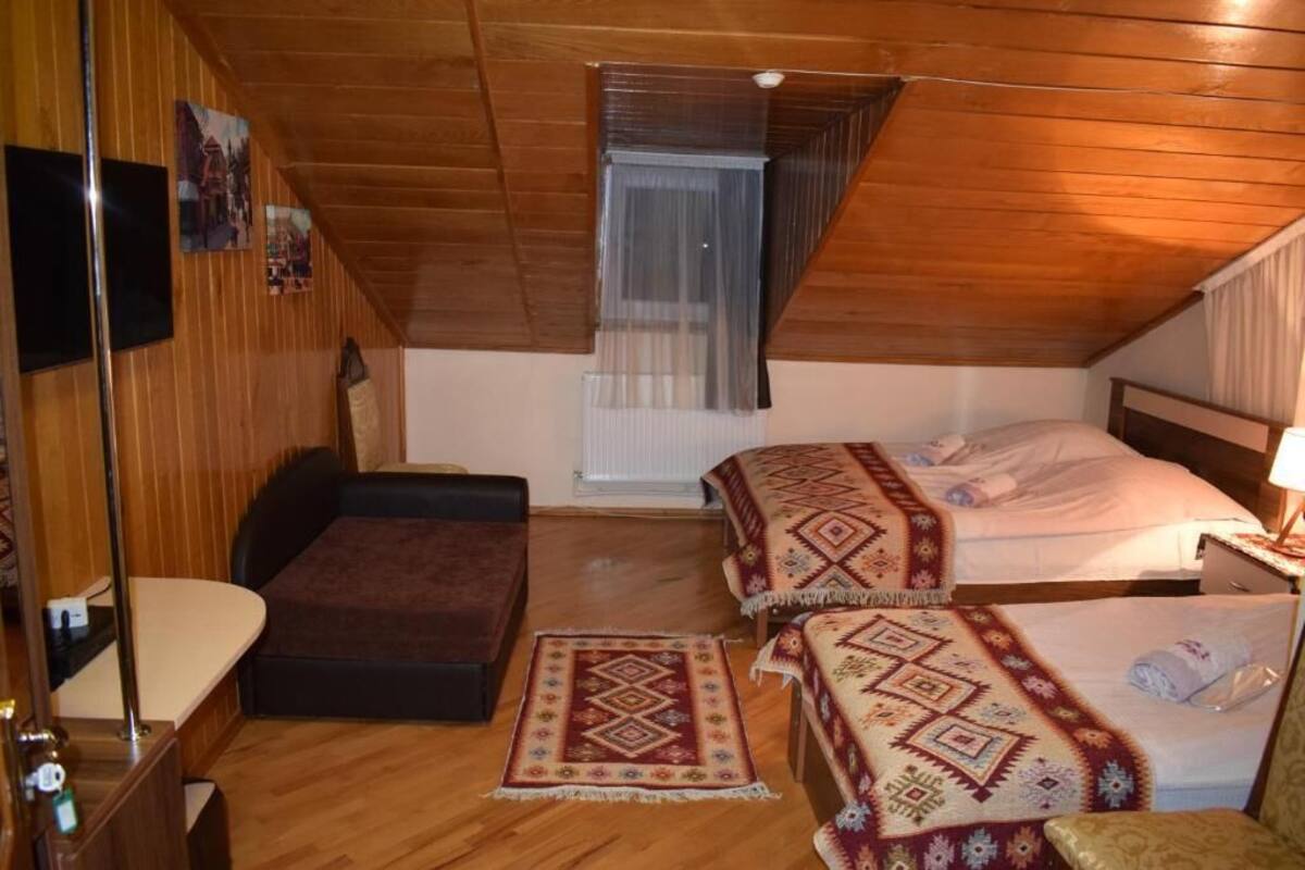 A small, cozy room with wooden walls and a sloped ceiling. There are two beds with patterned bedspreads, a bench, and a small table with a lamp.