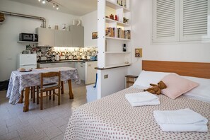 Un dormitorio con una cama, un osito de peluche, una mesita de noche, una silla y una kitchenette con una mesa, sillas y un microondas.