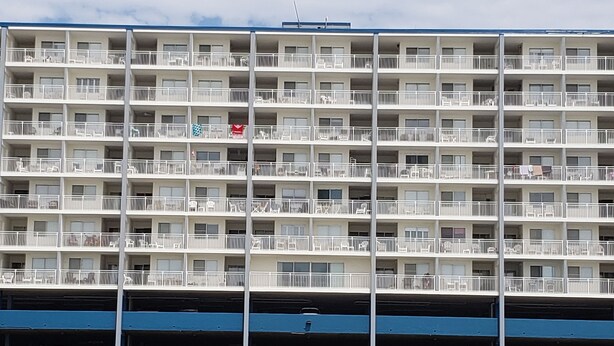 Ein mehrstöckiges Hotel mit Balkonen, einer Tiefgarage darunter und Strandliegen im Erdgeschoss.
